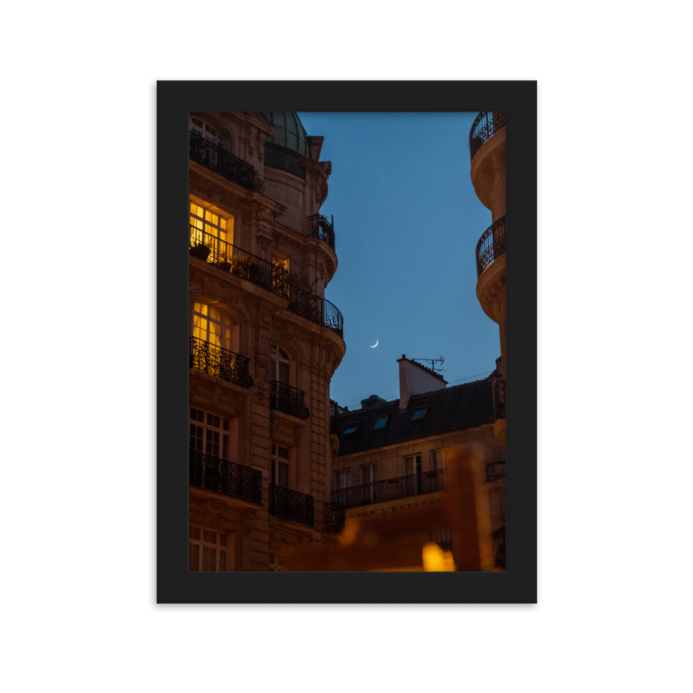 Croissant de lune entre des immeubles haussmanniens éclairés la nuit.