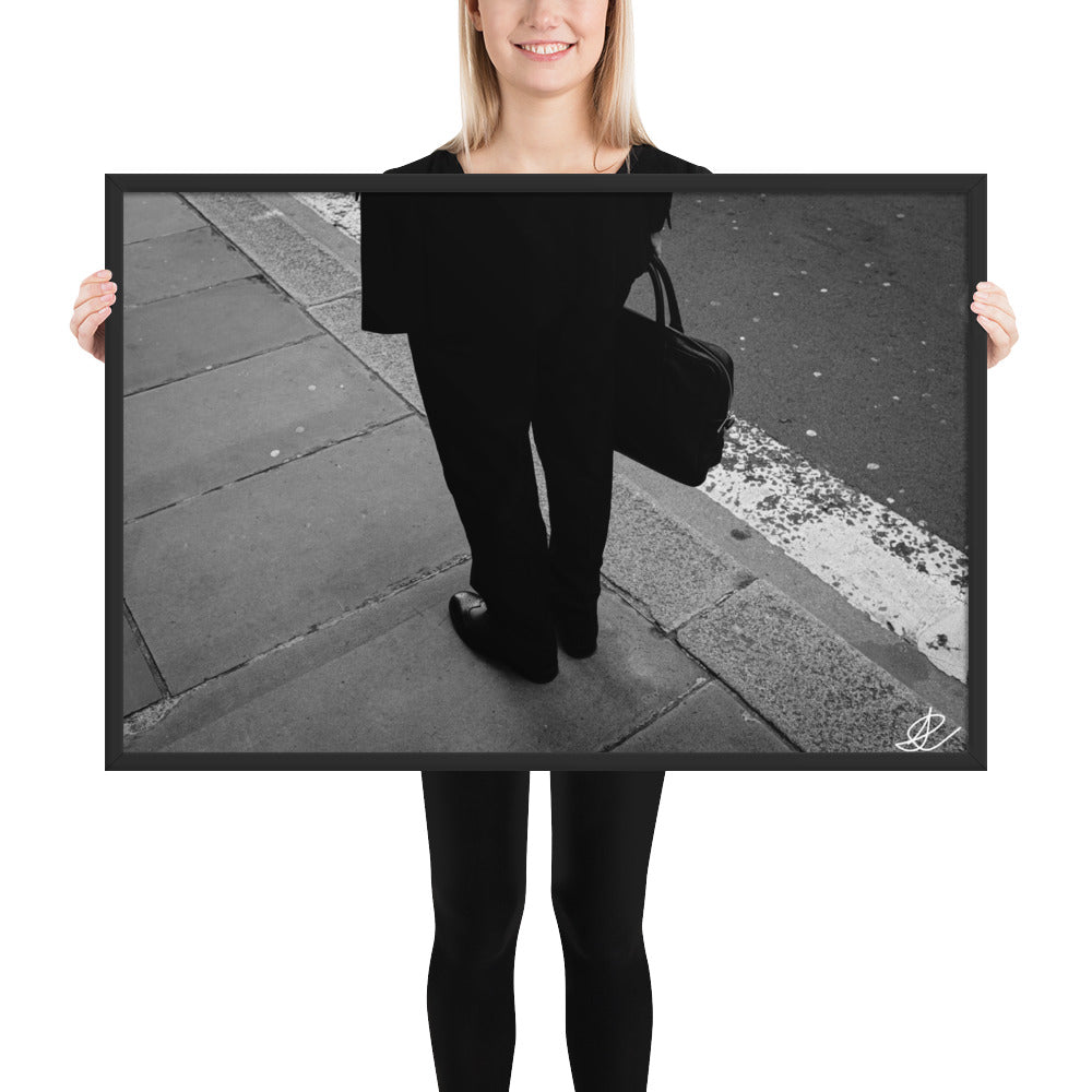 Photographie noir et blanc 'L'Homme d'Affaires' par Ilan Shoham, capturant une scène urbaine quotidienne avec un focus sur une paire de jambes, un sac à documents et un costume noir contre le décor brut du trottoir, encadrée élégamment pour un impact visuel puissant.