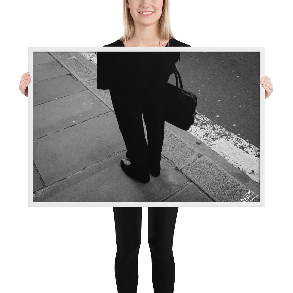 Photographie noir et blanc 'L'Homme d'Affaires' par Ilan Shoham, capturant une scène urbaine quotidienne avec un focus sur une paire de jambes, un sac à documents et un costume noir contre le décor brut du trottoir, encadrée élégamment pour un impact visuel puissant.