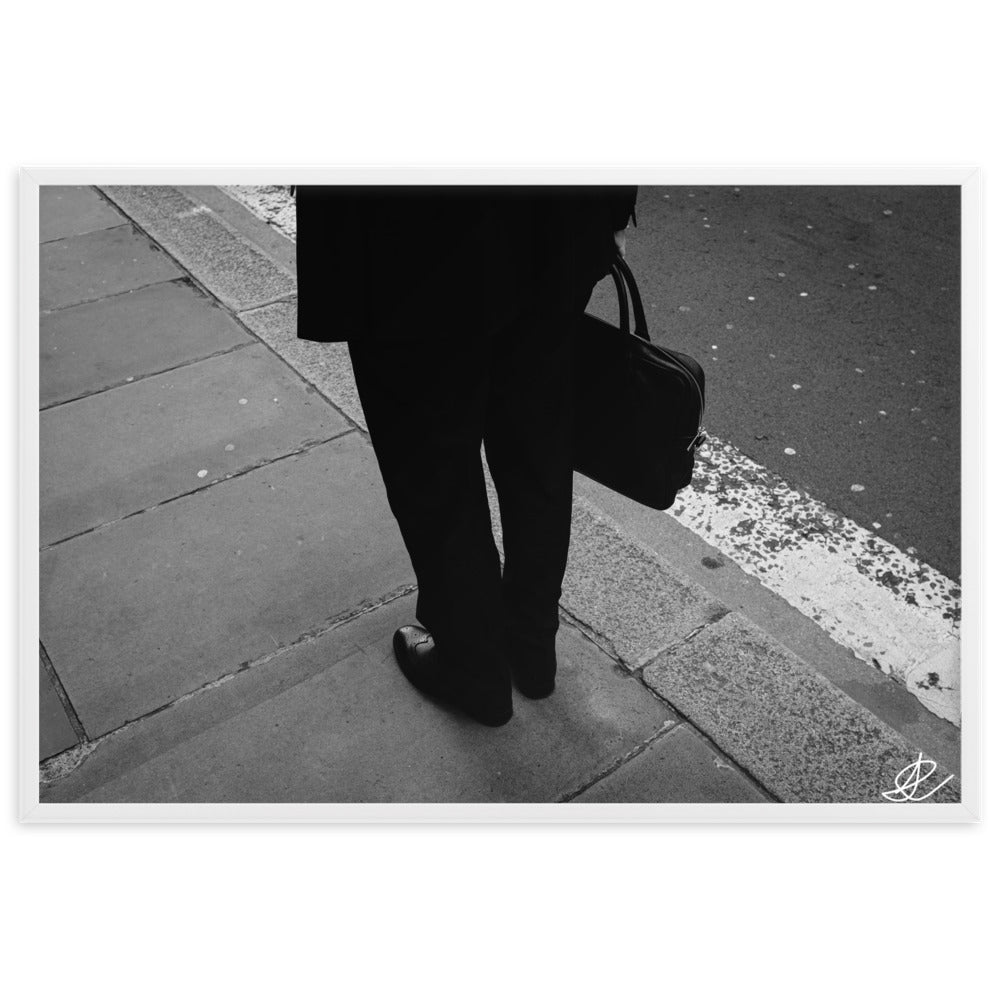 Photographie noir et blanc 'L'Homme d'Affaires' par Ilan Shoham, capturant une scène urbaine quotidienne avec un focus sur une paire de jambes, un sac à documents et un costume noir contre le décor brut du trottoir, encadrée élégamment pour un impact visuel puissant.