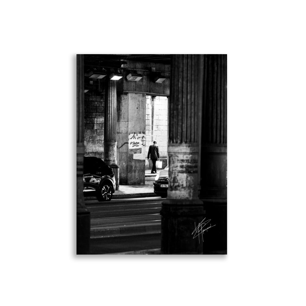 Photographie en noir et blanc, un homme sortant d'un tunnel sombre avec des sacs de courses, tandis que des voitures s'approchent de lui.