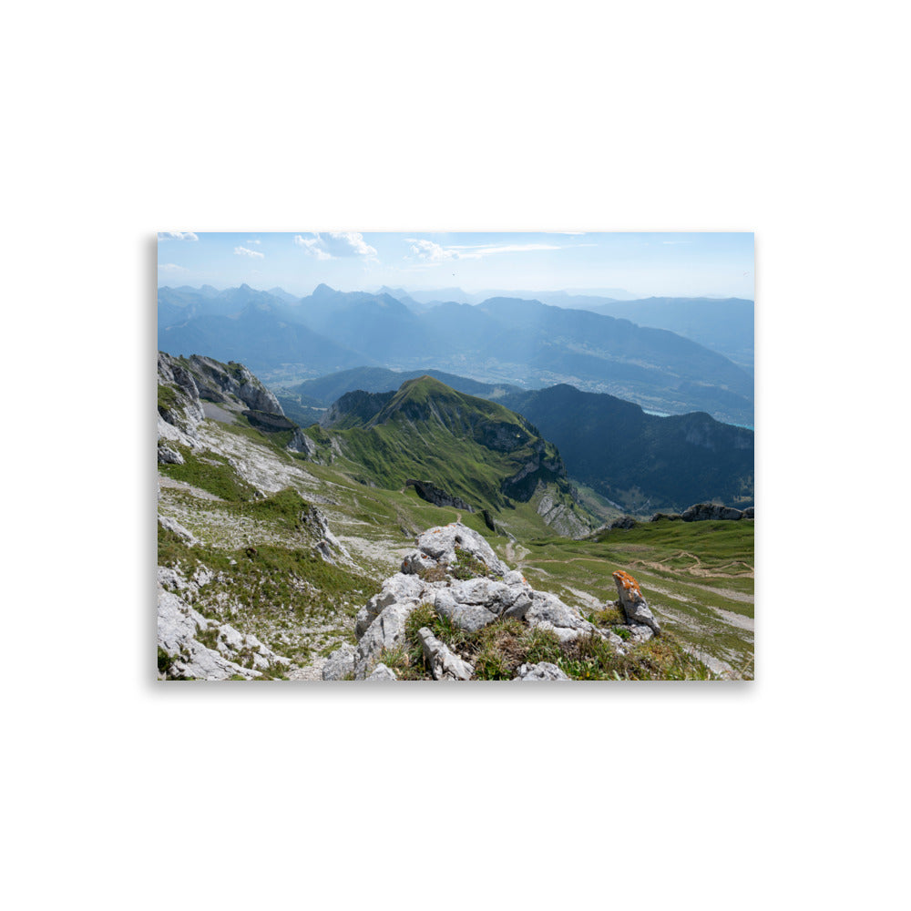 Vue panoramique du lac Annecy depuis les hauteurs, entouré de montagnes majestueuses avec un ciel serein au-dessus. Une représentation fidèle de la beauté naturelle des Alpes.