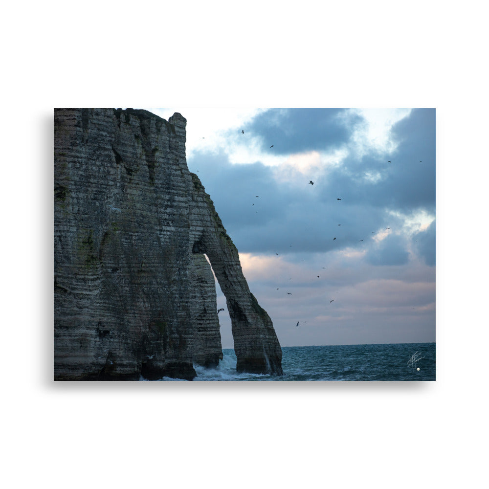 Vue panoramique des falaises d'Étretat avec des vagues s'écrasant puissamment sur la côte, sous un ciel nuageux où les mouettes virevoltent, incarnant la beauté sauvage de la Normandie.