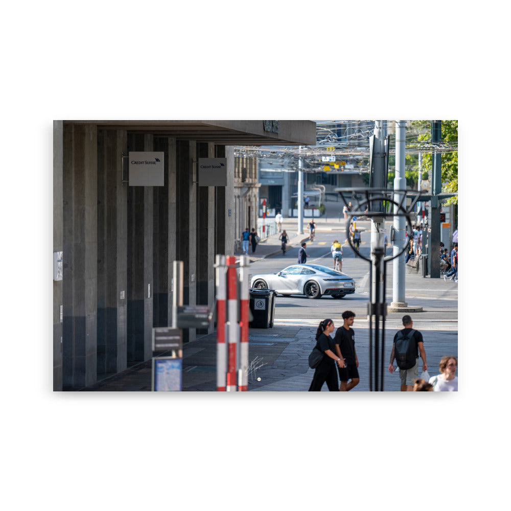 Photographie urbaine montrant la façade de la banque Crédit Suisse à Genève, avec des passants, des cyclistes, et une élégante Porsche 911 grise en mouvement au premier plan.