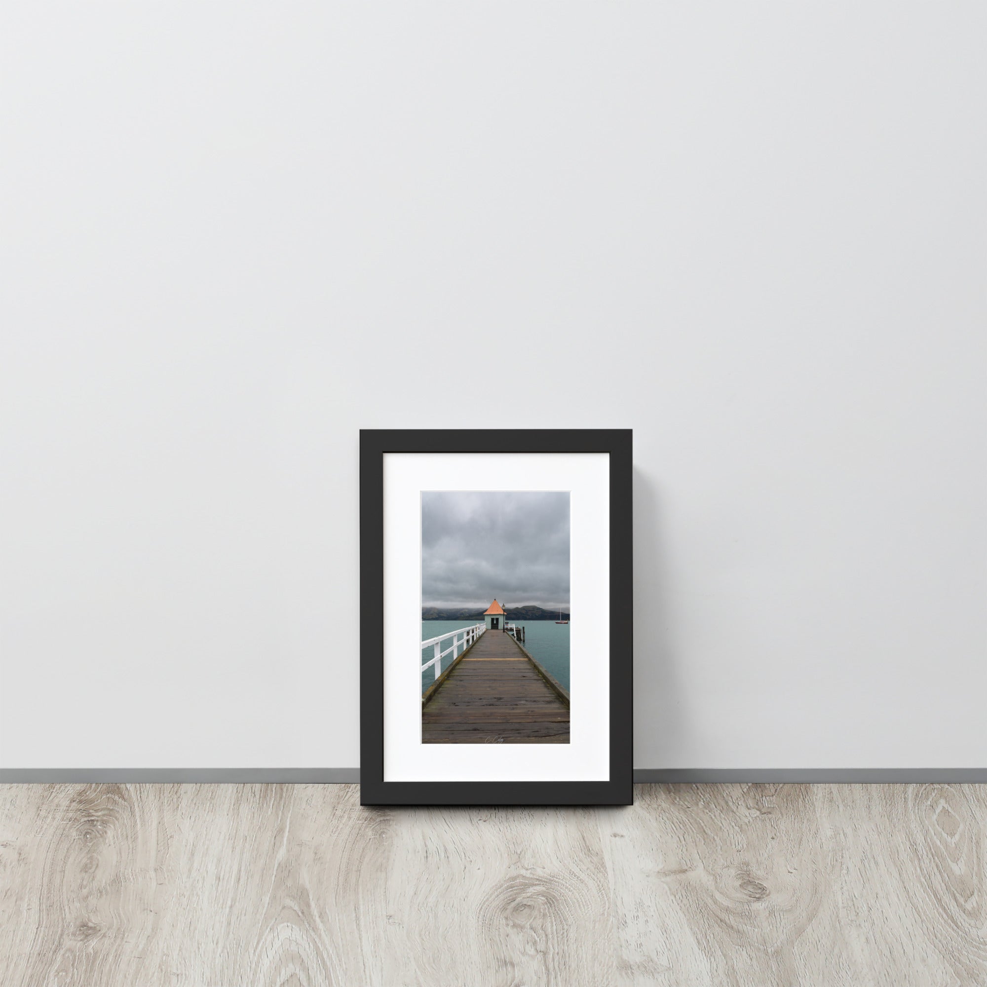 Photographie 'Akaroa NZ' de Charles Coley, offrant une vue sereine sur un lac néo-zélandais avec un petit abri au bout d'un ponton rustique, contrastant délicatement avec un arrière-plan de montagnes majestueuses et un ciel nuageux, ajoutant une dimension de tranquillité et d'évasion à votre espace.